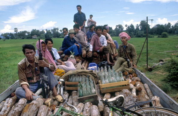 People sit on an open train car filled with timber. Some people are holding guns. There are other random items like a bike and bottles scattered around.