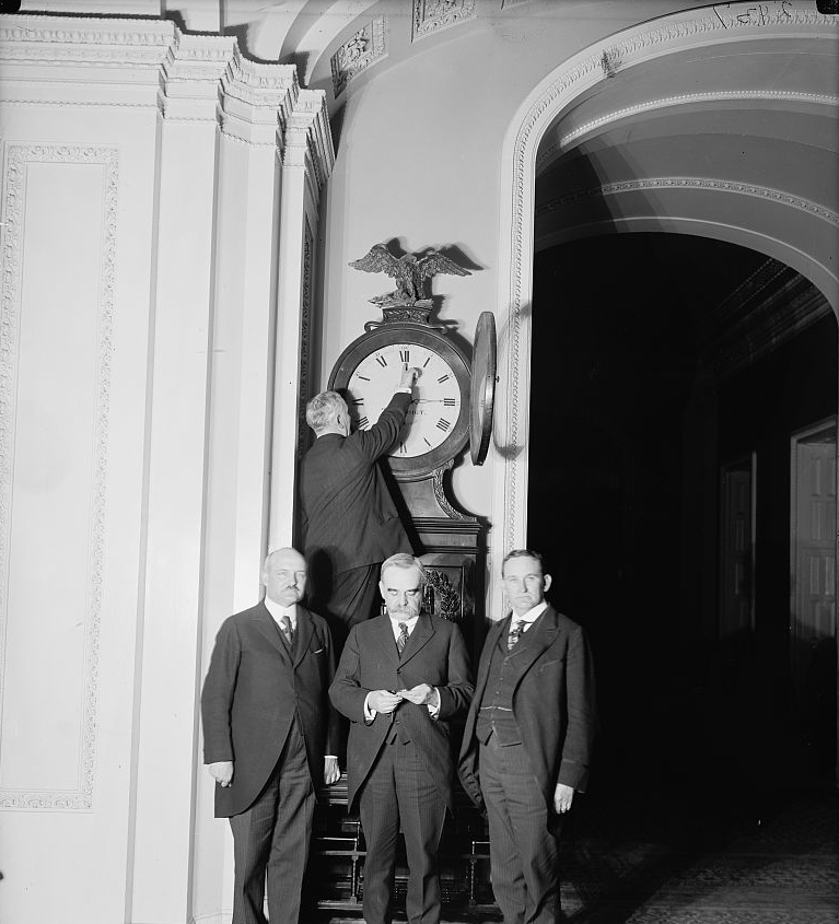 Three men in old-fashioned three-piece suits stand in front of a large grandfather clock as another man adjusts it.