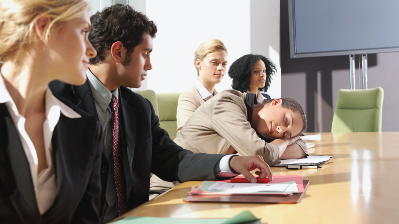 In this analogy for freeriding, a woman takes a nap at her desk during a business meeting while her co-workers.  