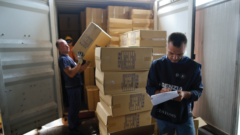 In this photo  customs officer inspects the contents of a shipping container at the harbor in Naples, Italy. Behind him a man unloads boxes from the shipping container.