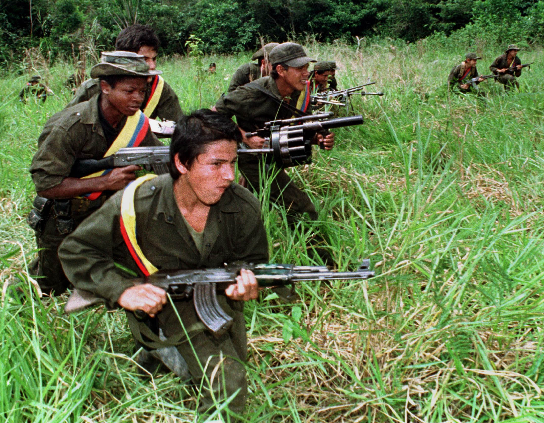 Guerrilla fighters of the Revolutionary Armed Forces of Colombia crouch in grass while patrolling the jungle with weapons.