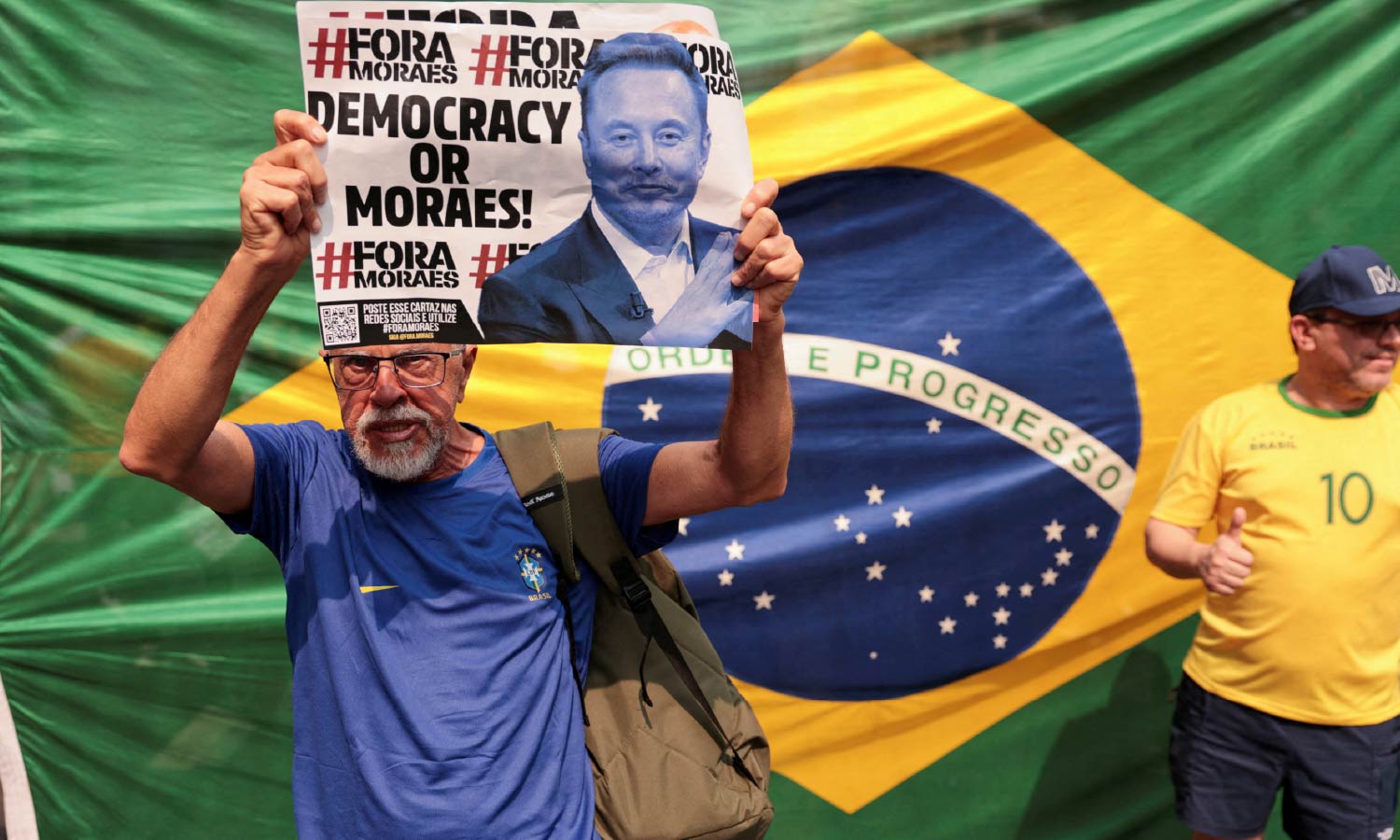 Bolsonaro supporter holding a sign protesting against judge Alexandre de Moraes. A large Brazilian flag is behind him.
