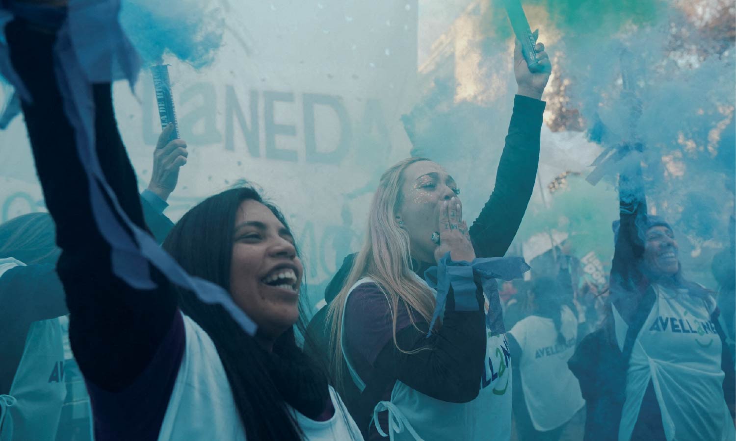 Two women at a protest for the "Ni Una Menos movement raise their hands while holding cannon smoke powder poppers as blue powder surrounds them.