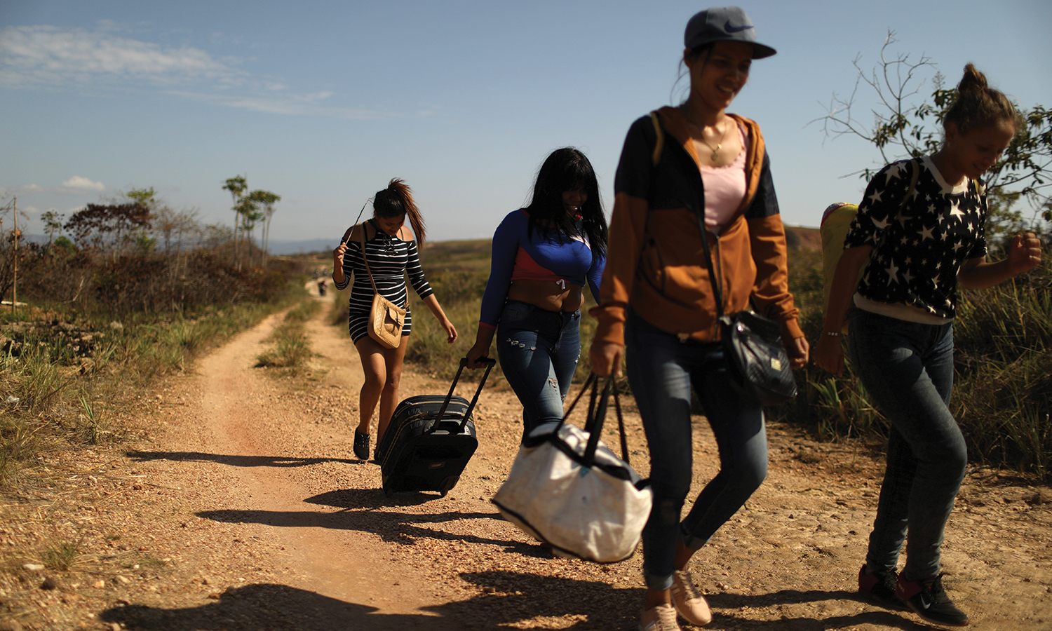 Woman carry bags while walking along dirt path.