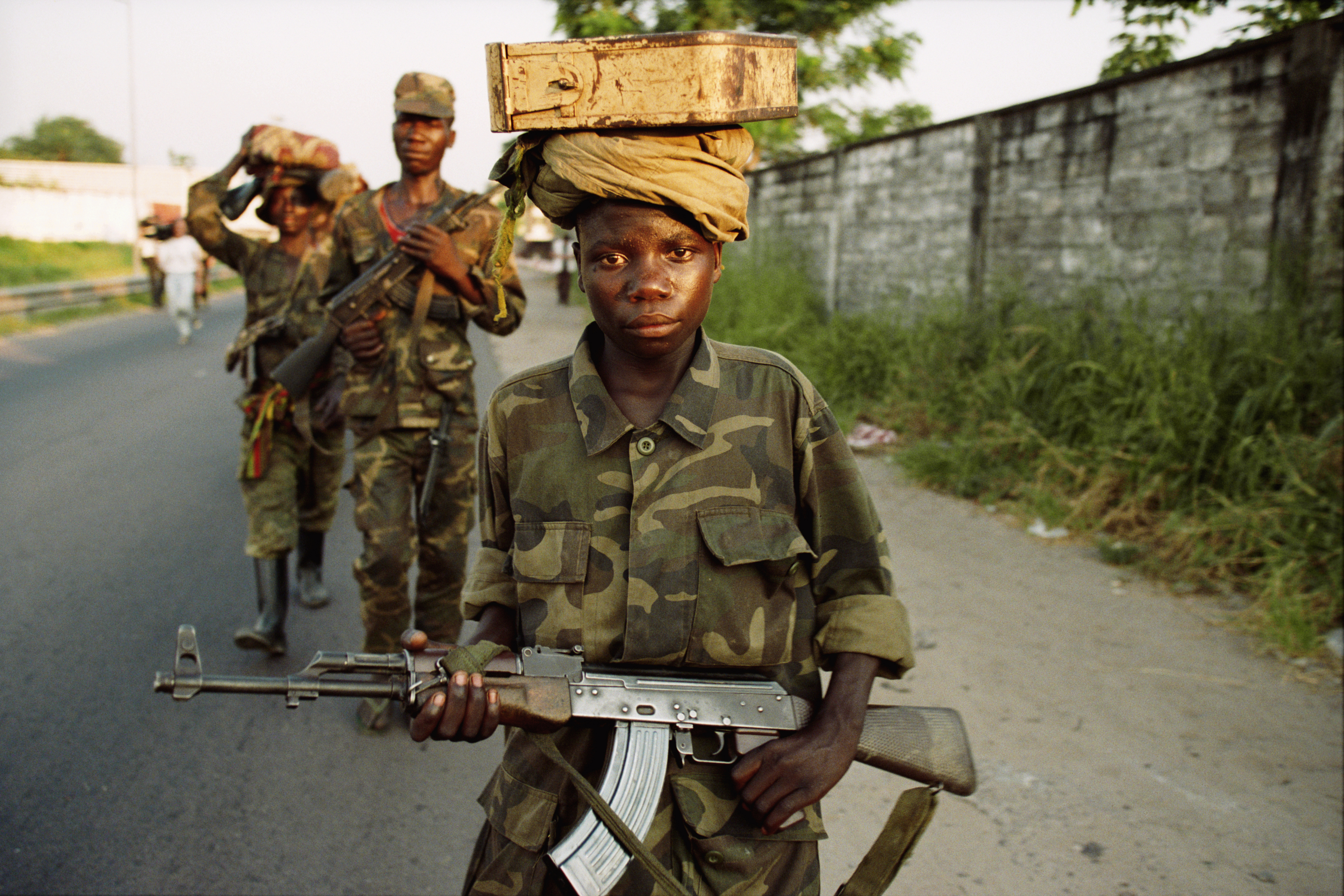 Child soldiers, wearing uniforms, hold guns and carry packages on their head.