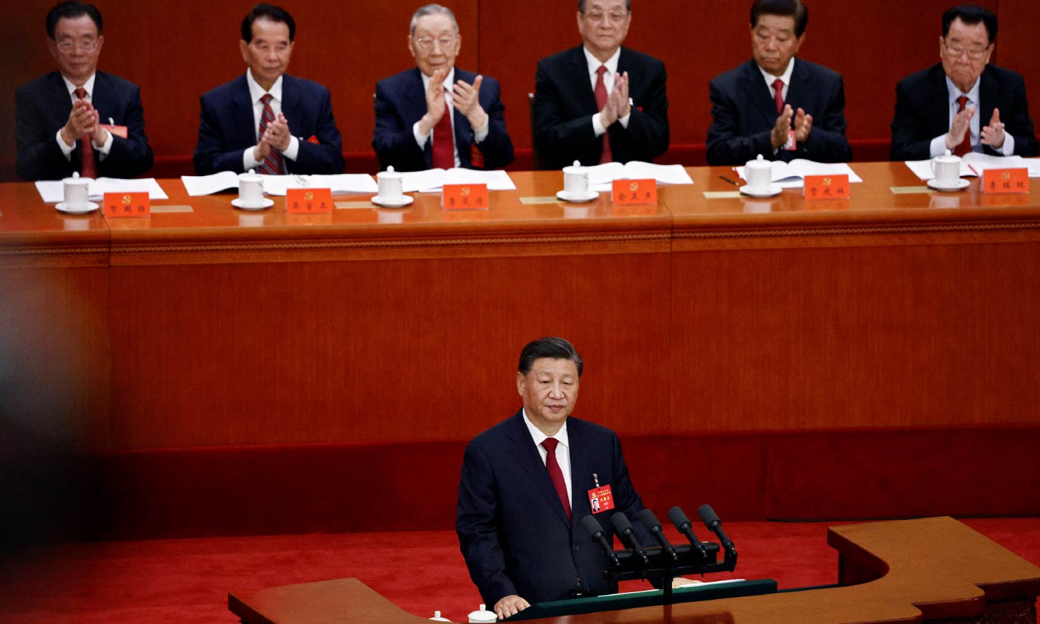 Chinese President Xi Jinping stands at a podium to give a speech. Other Chinese men sit behind him and applaud.