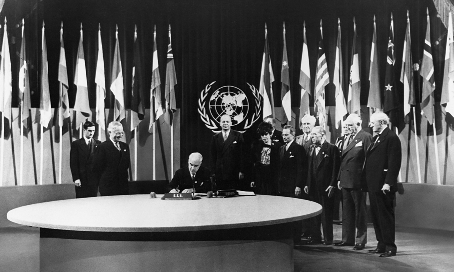 Edward Stettinius, seated at the table, signs the UN Charter while President Truman and others look on from behind him. Flags of UN nations surround the group.