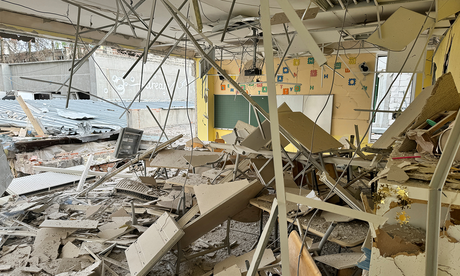 A view shows a destroyed school at the site of a Russian air strike. A chalkboard can be seen amidst the mess of destroyed desks, chairs, and walls.