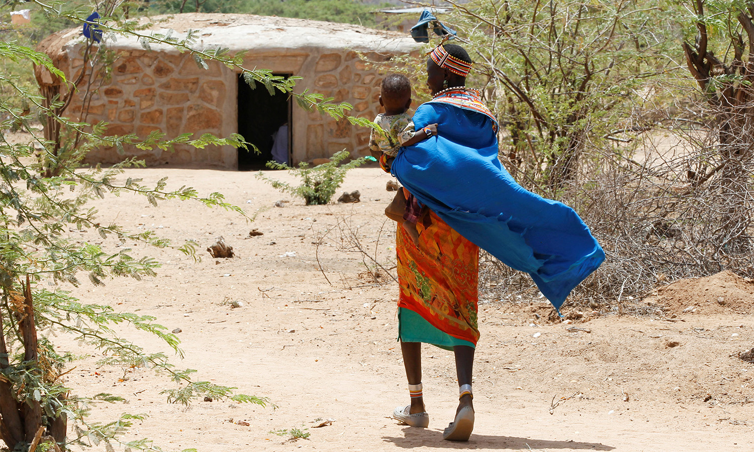A woman walks with a child as her clothing flows behind her. They are walking towards a mud dwelling surrounded by trees.