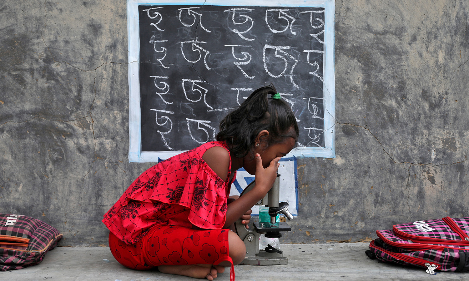A girl wearing a red outfit sits and uses a microscope outdoors. Behind her is a chalkboard with the alphabet.