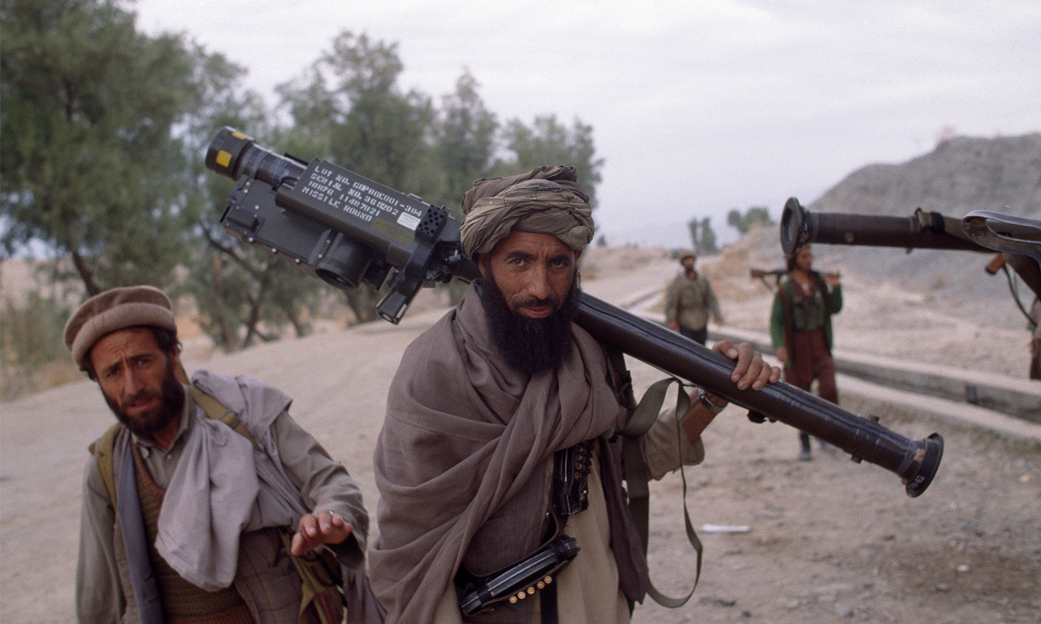 Bearded man in cloak and turban with several other men walks down dusty road while carrying stinger missile launcher over his shoulder.