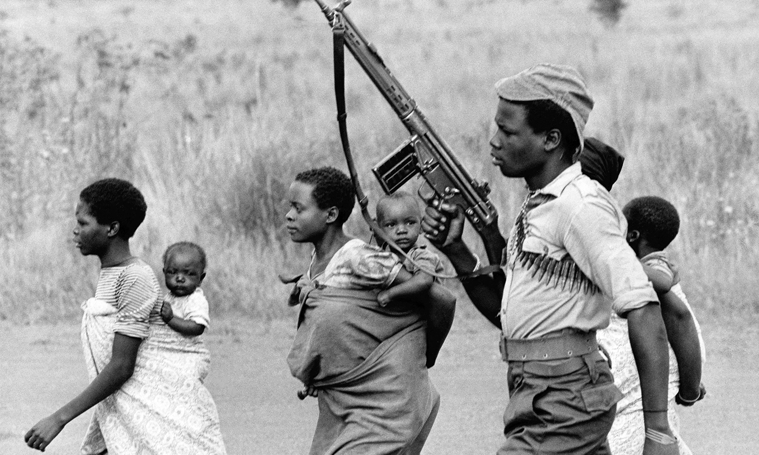 Black and white photo of man with ammo belt around his chest holding up a rifle as he walks with several women carrying young children.
