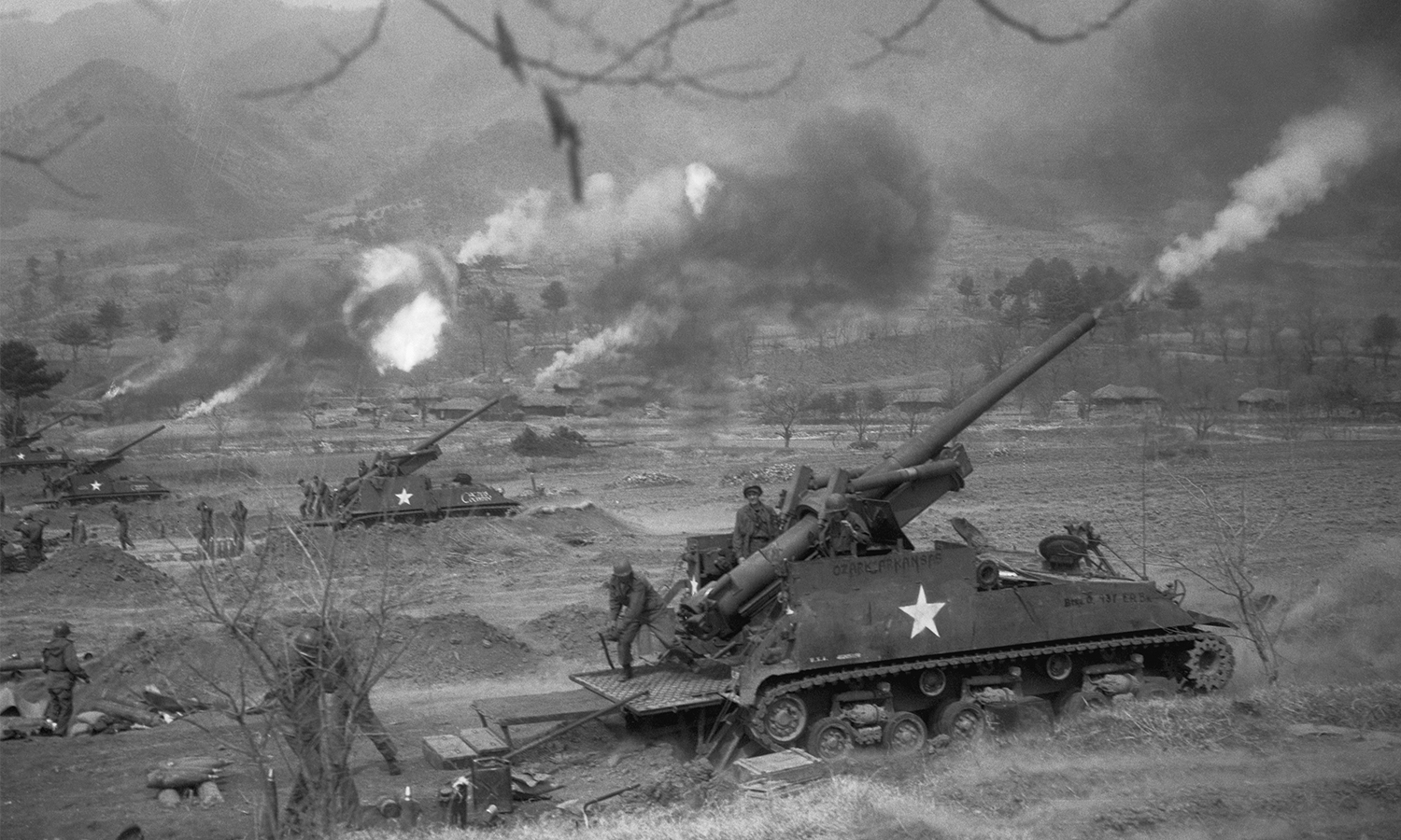 Large artillery with a star painted on their sides fire shells from a field with mountains in the distance.