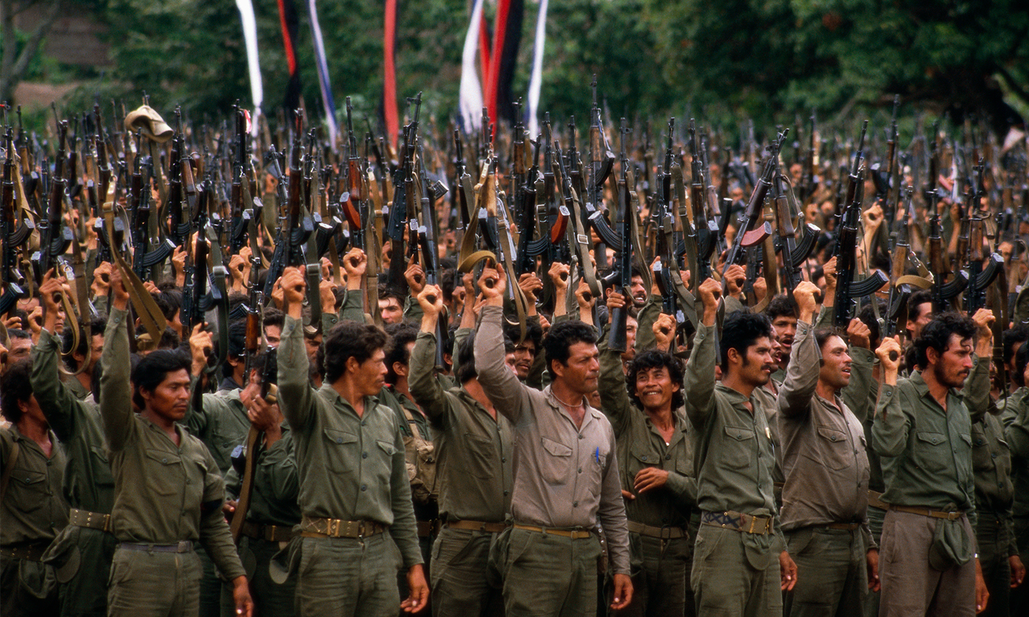 Rows of men in military fatigues raise rifles into the air with their right hands.