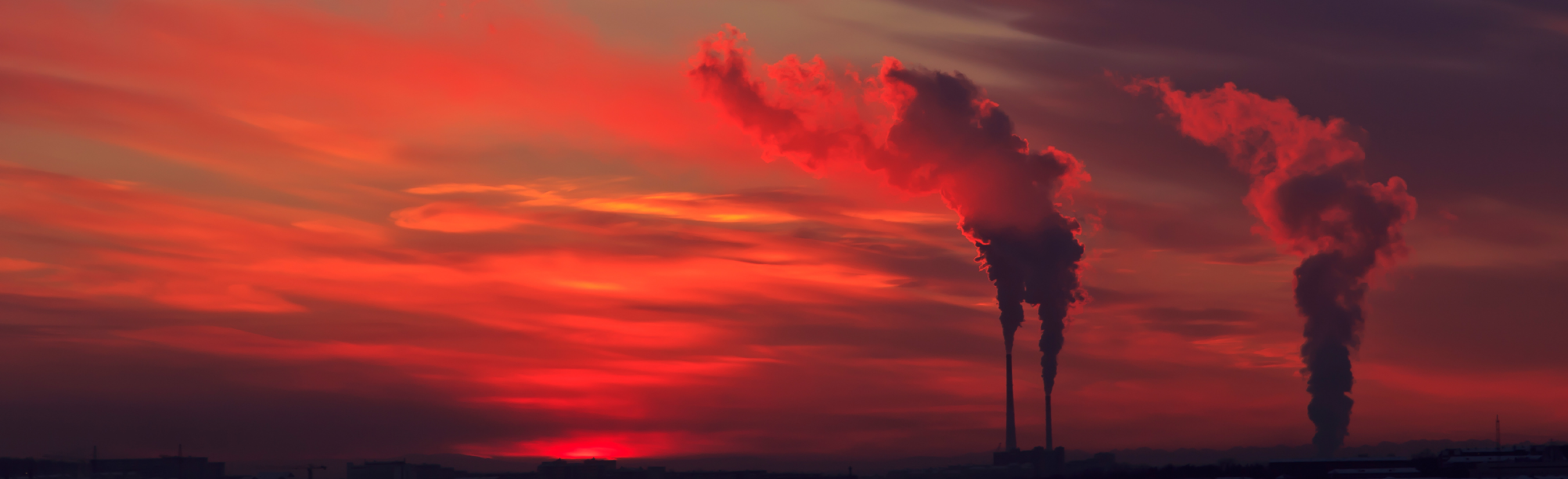 Red sky behind the silhouette of smokestacks emitting smoke.