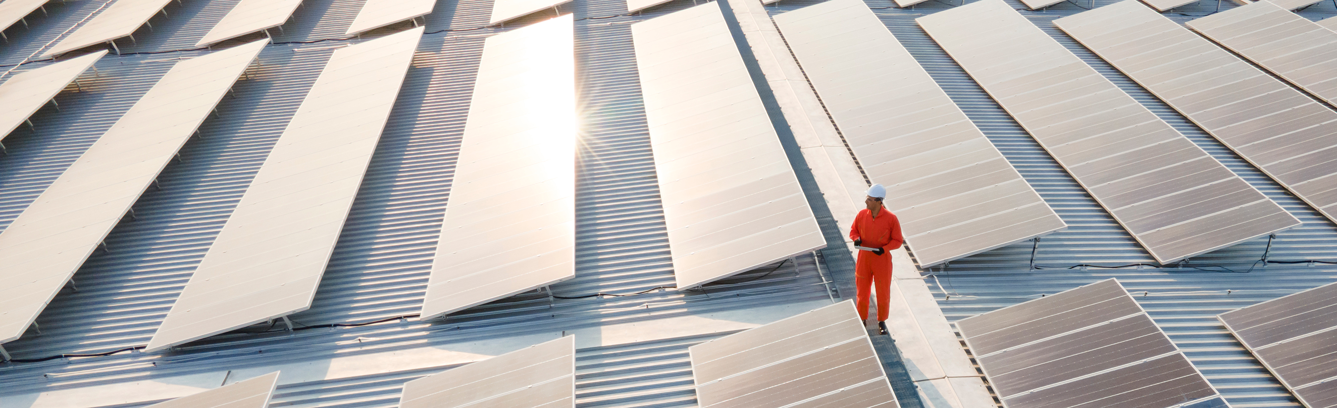 Engineer in orange coverall stands on top of roof covered in rows of solar panels shining in the sun.