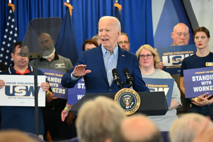 President Joe Biden speaks at a podium with united steelworkers union members holding union signs behind him.