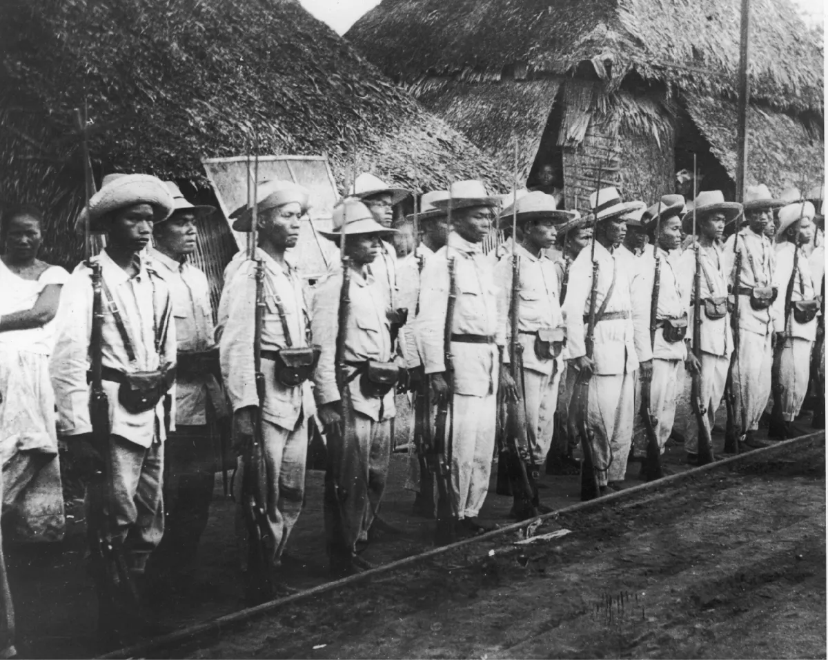 Philippine rebels from the Philippine-American War stand in a line holding rifles with bayonets.