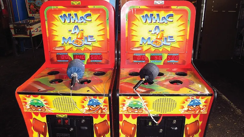 A picture of two Whac-A-Mole games at an arcade. In this game  a player uses a soft hammer to hit plastic moles that randomly pop up from holes in a cabinet. Each time a mole is knocked down, another appears in a new hole. 