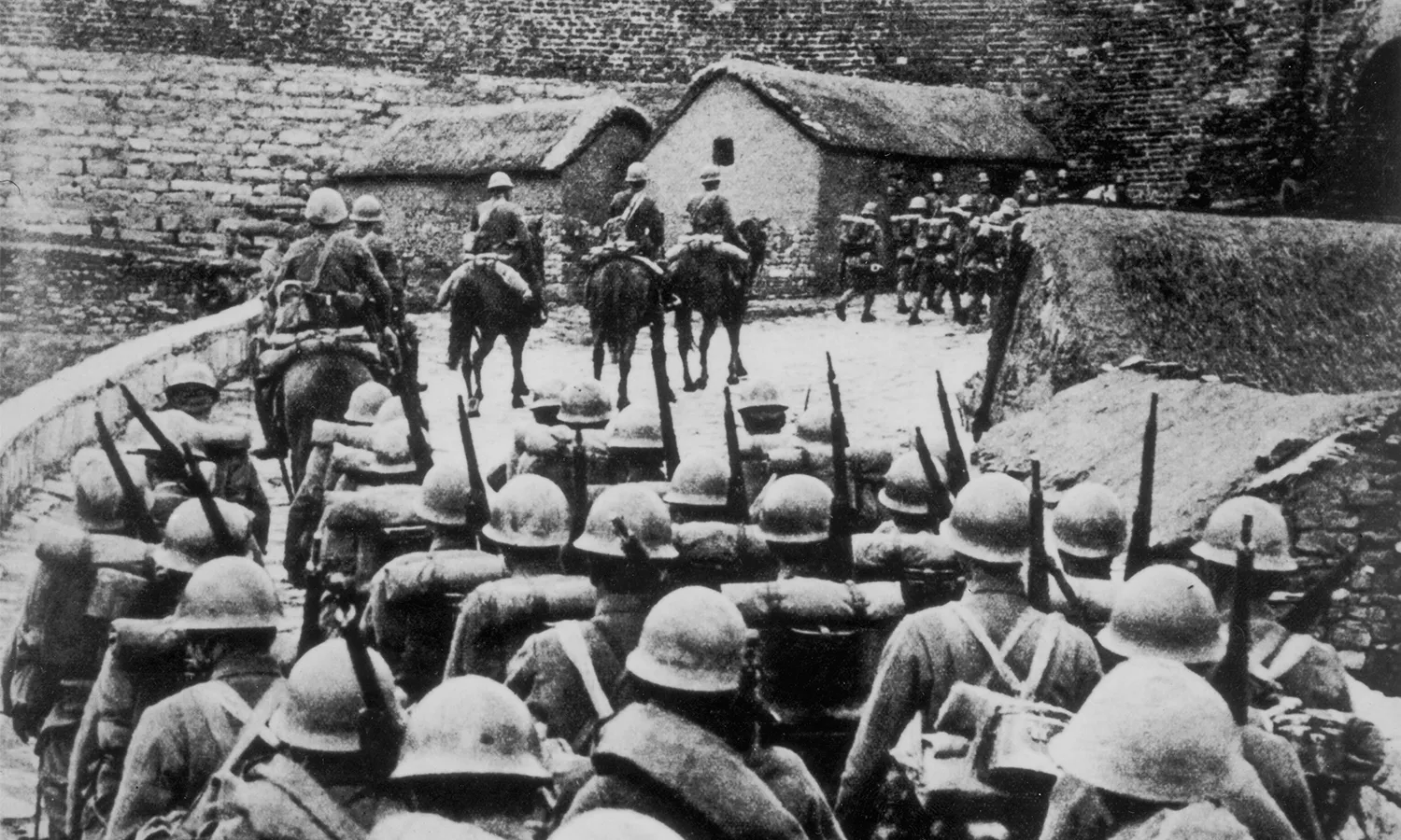 A black and white photo of armed Japanese troops on horseback and on foot entering Manchuria. The soldiers are walking towards a tall brick wall.