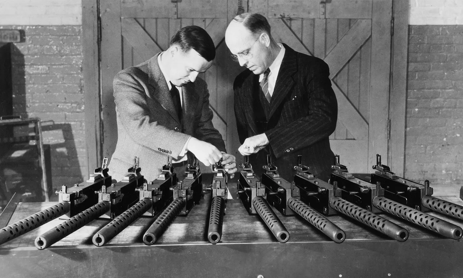 A black and white photo of two men inspecting machine guns laid out on a table.