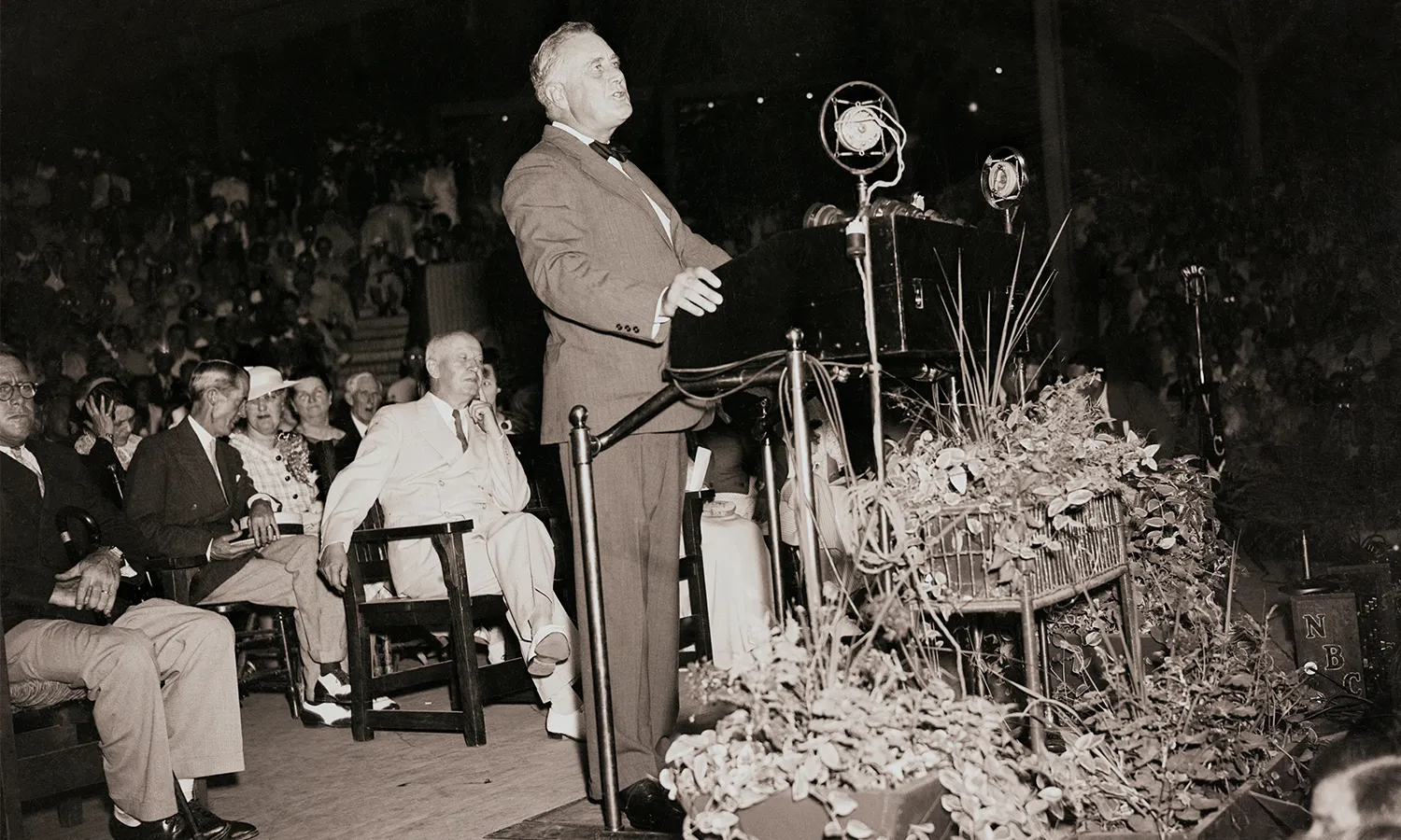 A black and white photograph of President Franklin D. Roosevelt standing at a podium delivering his "I Hate War" speech at Chautauqua Institution in Chautauqua, New York on August 14, 1936 as audience members sit behind him..