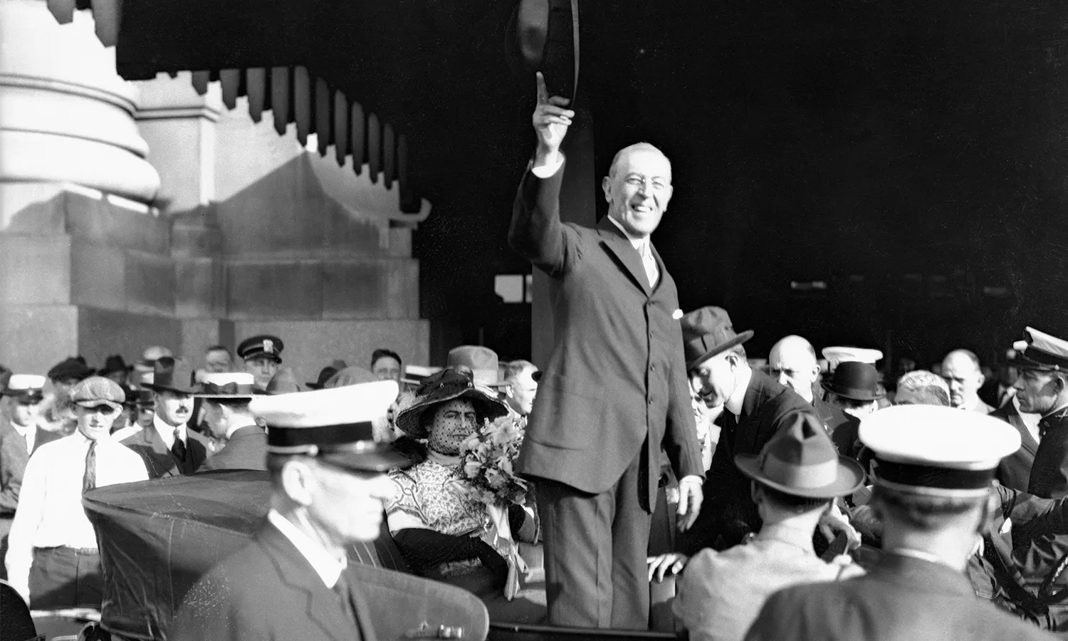 A black and white image of President Woodrow Wilson standing up in a car and raising his hat to the public while smiling.