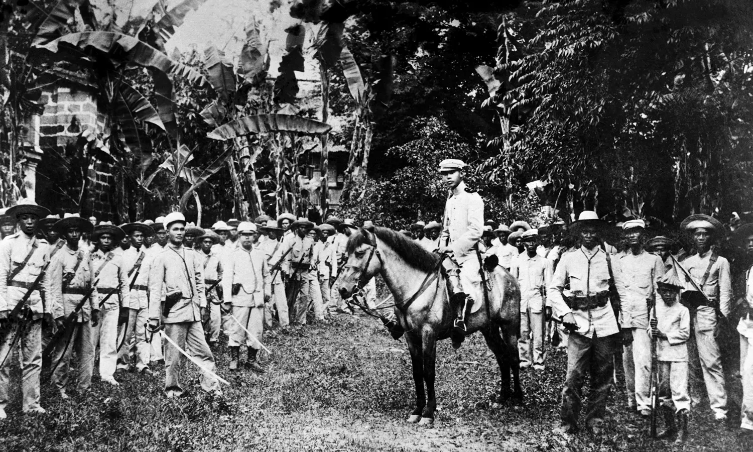 Black and white photograph of General Emilio Aguinaldo and his army. Aguinaldo is atop a horse as his army stands around him with weapons in their hands.