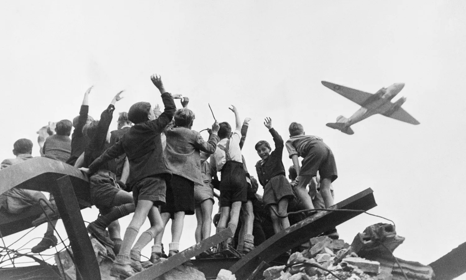 A black and white photo of children climbing rubble to wave at a plane.