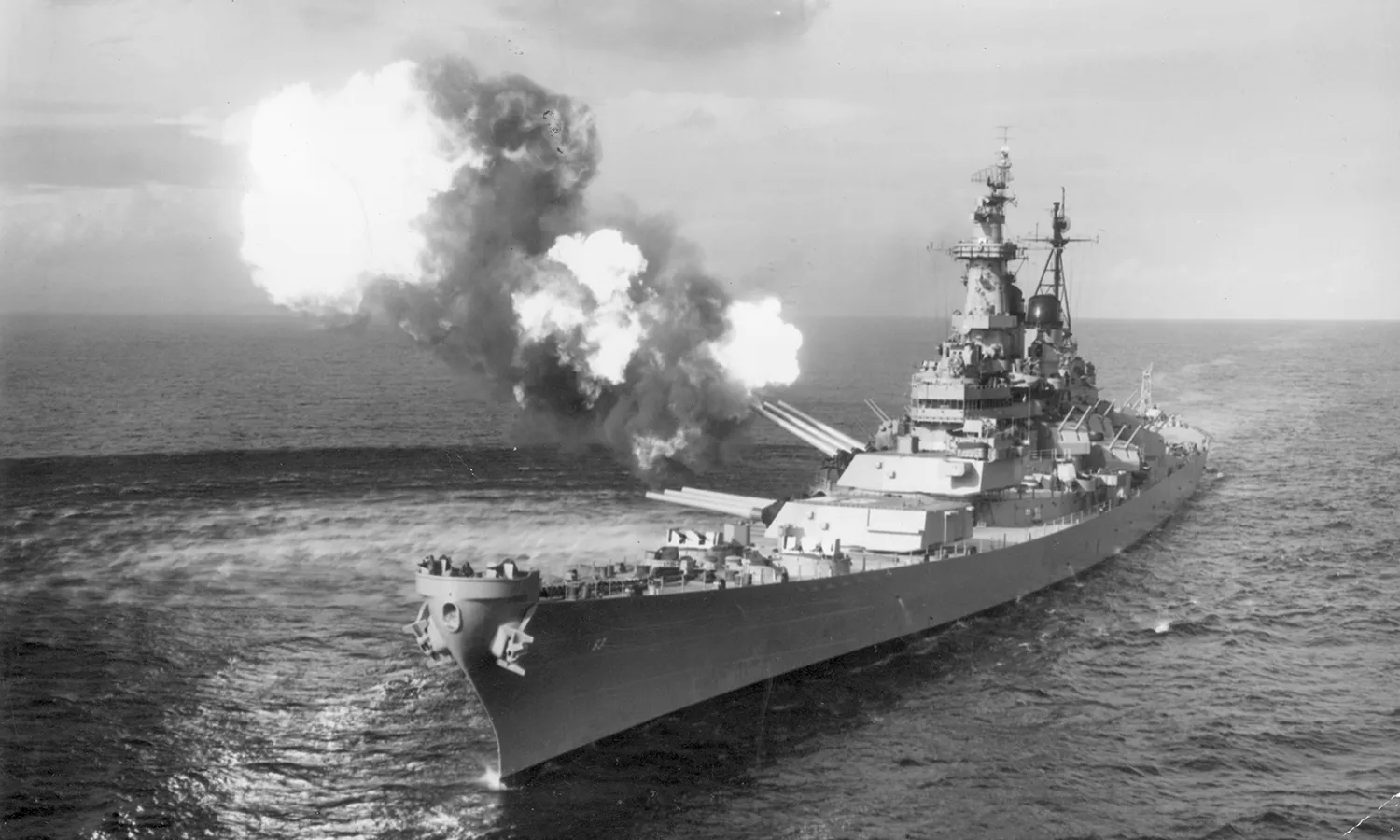 A black and white photo of a battleship in the ocean firing towards the left. 