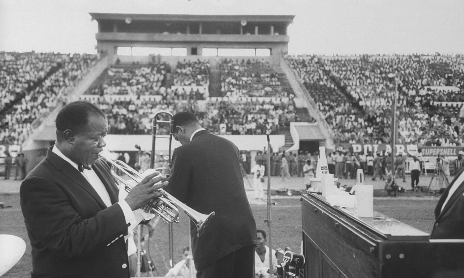 A black and white photograph of Louis Armstrong playing the trumpet in front of a large crowd gathered at a stadium.