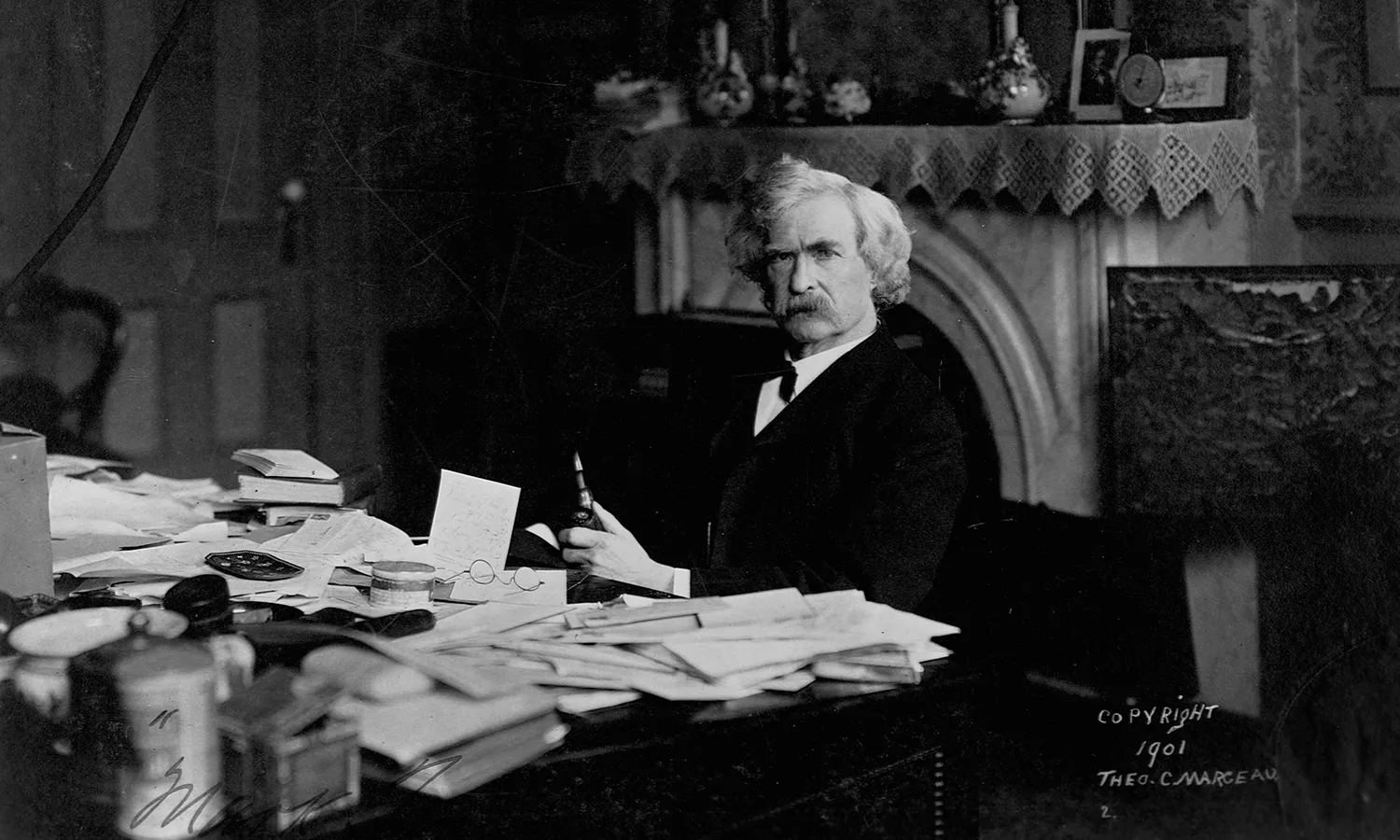 A black and white photo of Mark Twain seated at his desk with papers strewn all over his desk. 