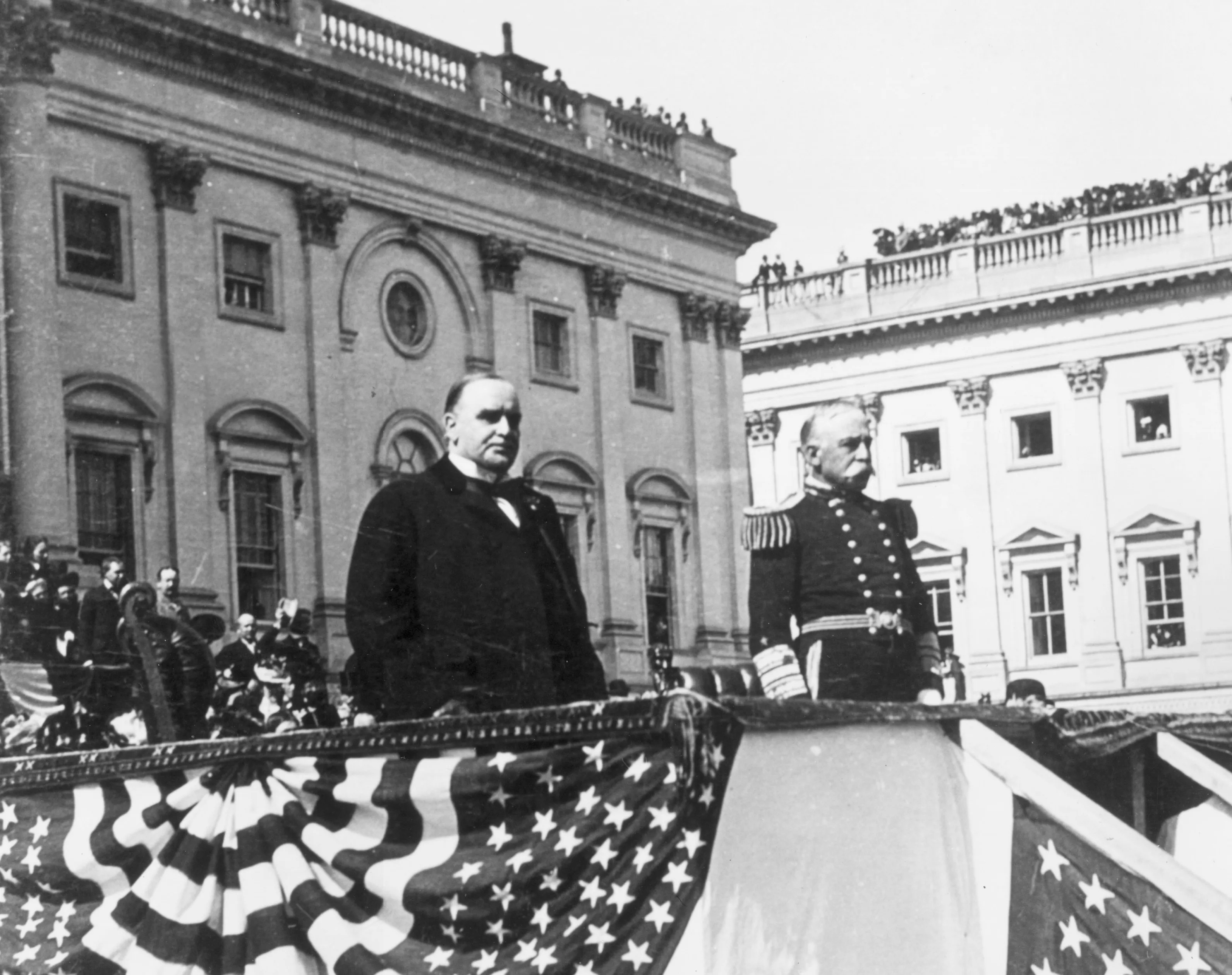 A black and white photograph showing President McKinley and Admiral Dewey, in uniform, standing on a podium decorated with American flags looking at an audience.