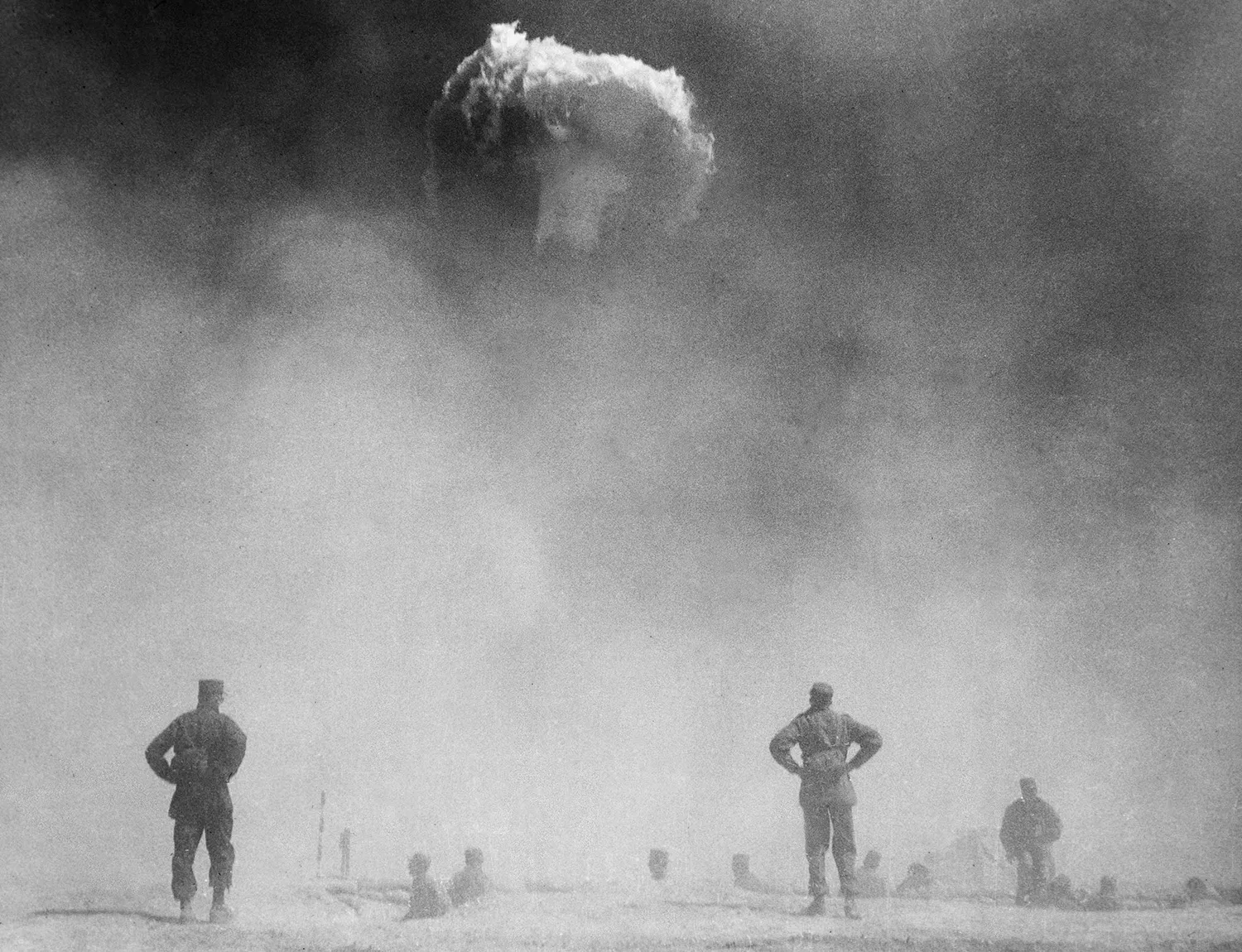 A black and white photograph of a U.S. nuclear test. Multiple people stand at a distance and watch a mushroom cloud rise as smoke and dust fill the air.
