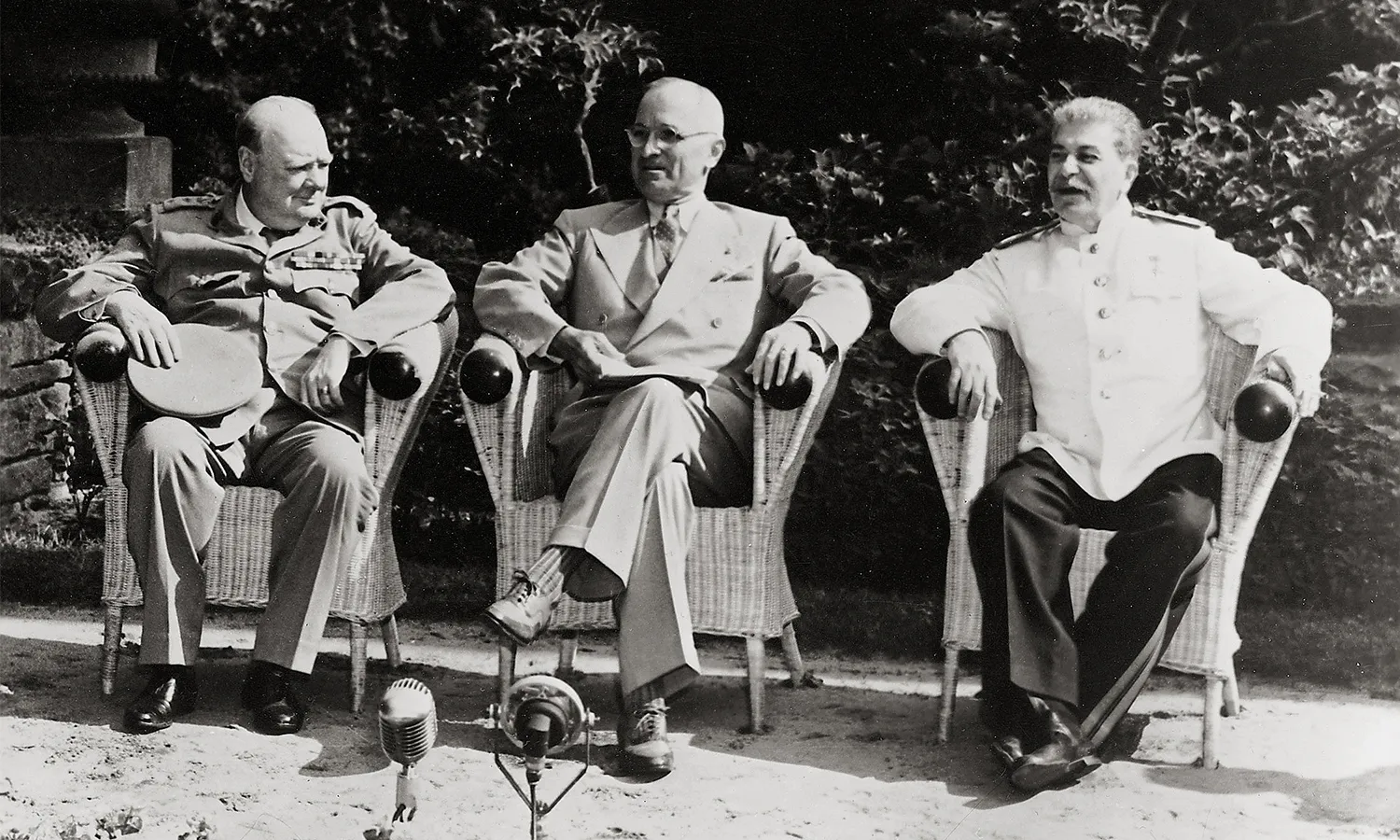 A black and white photograph of Winston Churchill, Harry S. Truman, and Joseph Stalin from left to right. All three men are seated on chairs outside and two mics are placed before them.