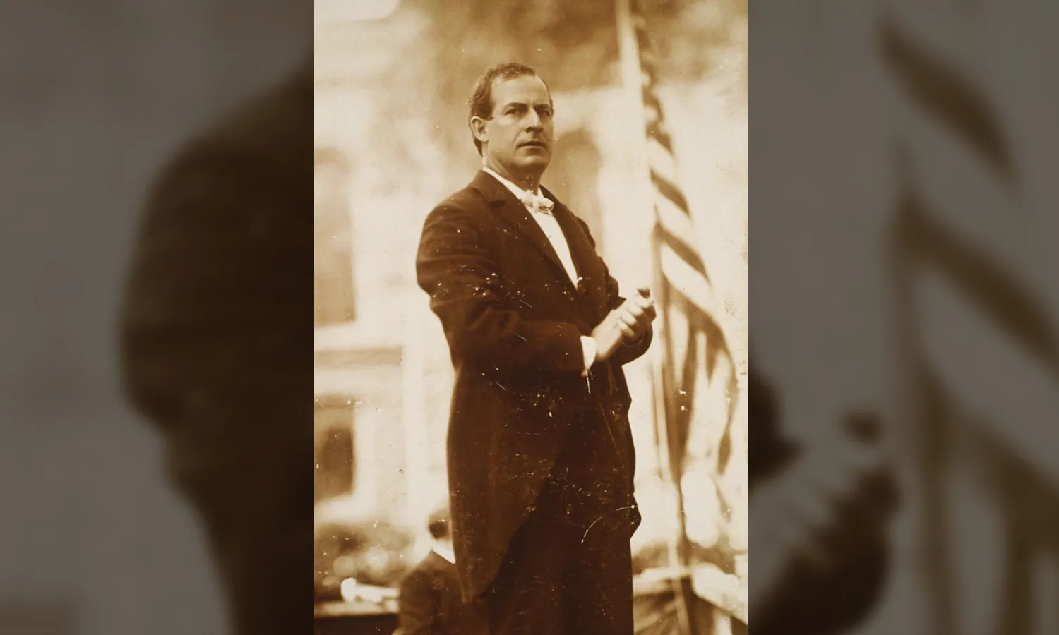 A sepia colored photograph of William Jennings Bryan standing in a suit next to the American flag.